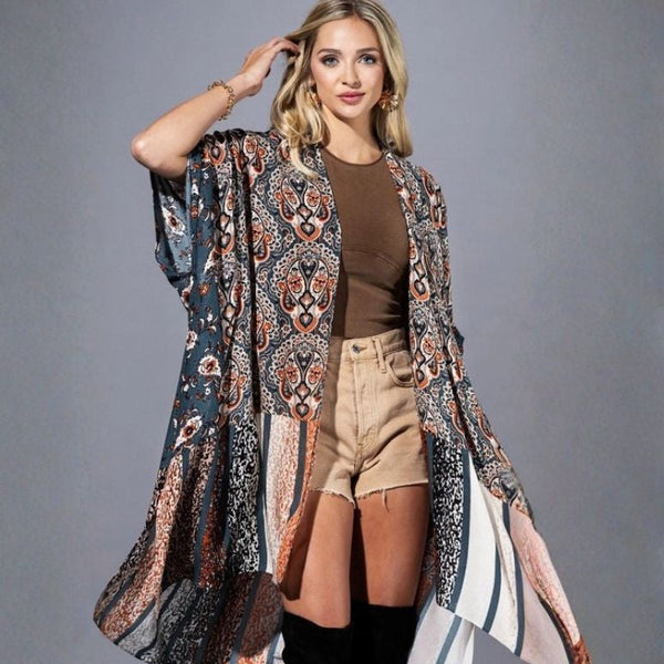 A woman models a long, open-front kimono-style cardigan with a multicolor paisley and patchwork print. The cardigan features a mix of dark blue, orange, beige, and white patterns.