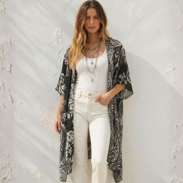 A woman stands against a textured white wall wearing a black and white patterned open-front kimono cardigan over a white tank top and white pants.