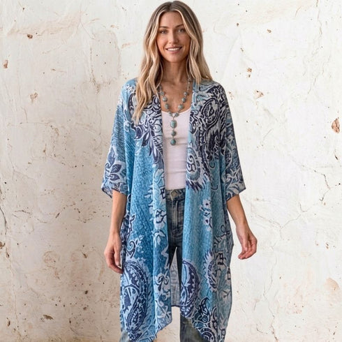 A woman stands against a textured off-white wall wearing a lightweight open-front Soulza kimono cardigan with a blue and navy paisley pattern over a white top and jeans.