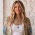 A woman with long blonde hair wearing a white ribbed tank top, a colorful floral-patterned kimono, and layered silver boho necklaces with turquoise stones, standing against a textured off-white wall.
