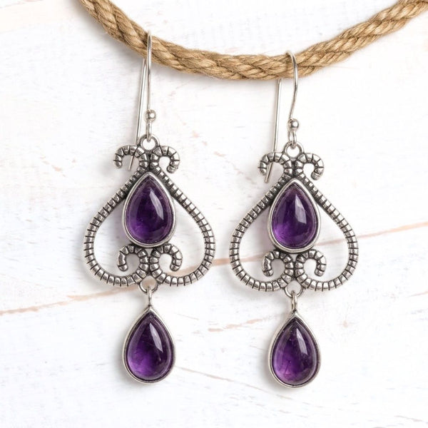 A pair of silver-toned teardrop dangle earrings with purple amethyst gemstones, hanging from a natural jute rope against a white textured background.