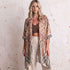 A woman stands against a textured off-white wall wearing a lightweight open-front kimono cardigan with a paisley print in beige, brown, and muted blue-gray tones over a brown ribbed crop top and cream pants.