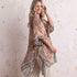 A woman wearing a lightweight open-front kaftan-style cover-up with a paisley print in beige, brown, muted green, and soft blue tones, standing against a textured light beige wall.