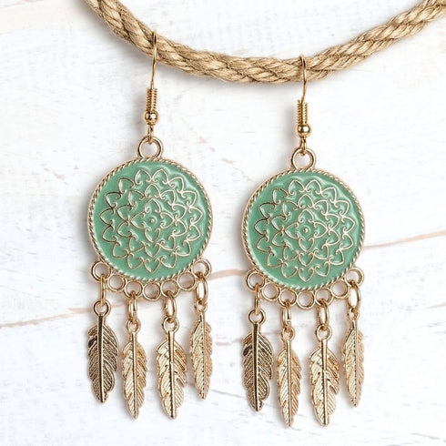 A pair of bohemian-style dangle earrings with mint green circular enamel inserts featuring gold filigree and hanging gold-toned feathers, displayed on a white textured background with a braided rope prop.
