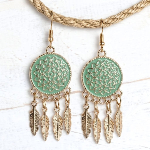 A pair of bohemian-style dangle earrings with mint green circular enamel inserts featuring gold filigree and hanging gold-toned feathers, displayed on a white textured background with a braided rope prop.