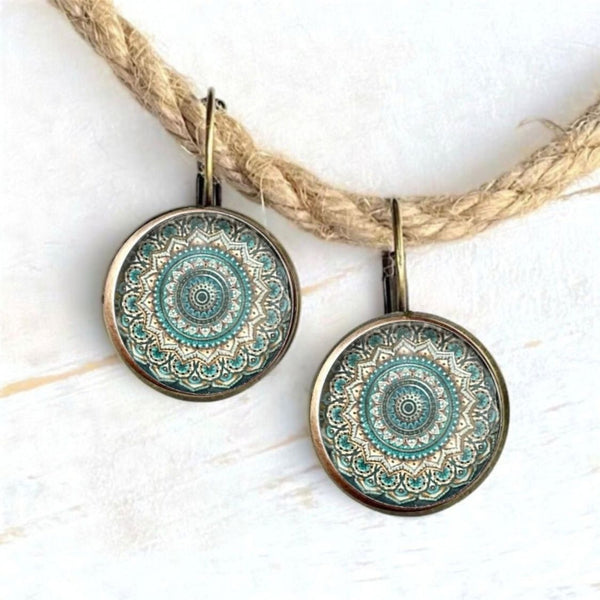 A pair of round drop earrings with intricate teal and gold mandala patterns inside antique bronze-toned bezels, hanging from matching hooks against a white background with a decorative jute rope.