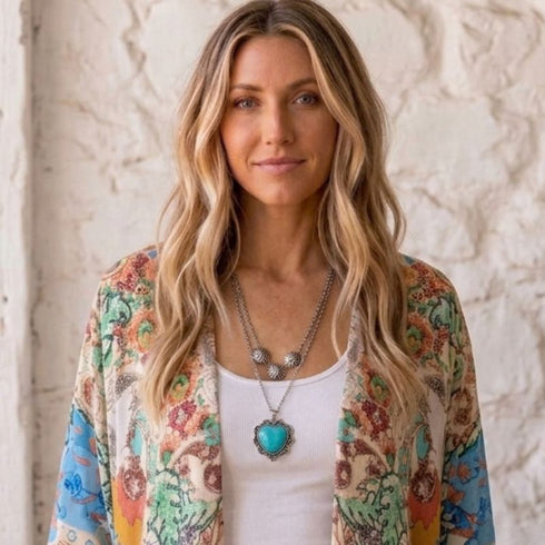 A woman with long wavy blonde hair is shown from the chest up, wearing a white tank top under a lightweight open-front cardigan with a colorful floral and botanical print. She is also wearing layered silver necklaces, including one with a large turquoise heart pendant.