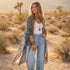 A woman stands in a desert landscape wearing a long sheer patterned kimono cardigan over a white tank top and light blue jeans. The cardigan features a navy blue base with intricate orange, beige, and gold floral and geometric patterns.