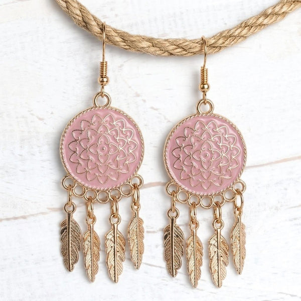 A pair of bohemian-style dangle earrings with pink circular enamel medallions featuring gold filigree detailing, hanging from gold-toned hooks and dangling gold-toned feather charms, displayed on a braided gold necklace over a white wood background.