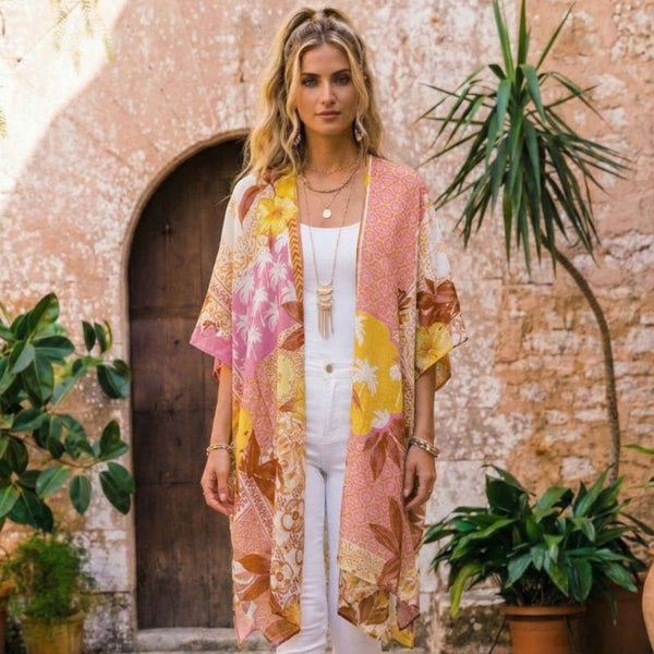 A woman stands outdoors in front of a rustic plaster wall and wooden door, wearing a lightweight open-front floral kimono cardigan over a white outfit. The cardigan has a warm pink base with large yellow, peach, and brown floral and botanical print areas.