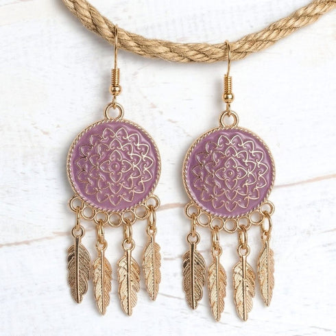 A pair of bohemian-style dangle earrings with lavender purple circular medallions featuring gold filigree patterns, hanging from gold-toned fishhook wires with dangling gold feathers, displayed on a white wood background.