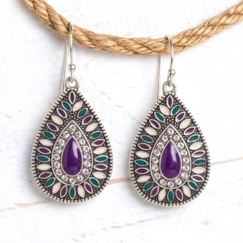 A pair of teardrop-shaped silver drop earrings with purple teardrop center stones and surrounding silver, white, purple, and teal enamel details, hanging against a light wood background with a rope prop.