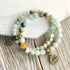 Two beaded bracelets with natural stone beads and bronze-toned charms, displayed on a rustic wooden surface next to a white painted wood block.