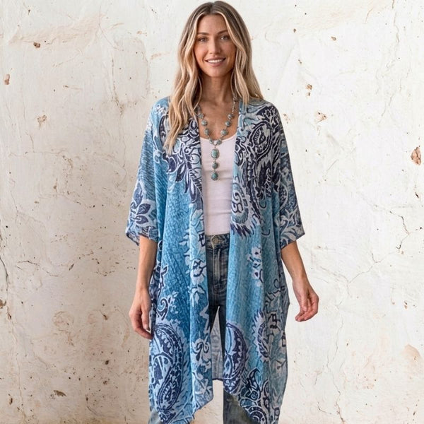 A woman stands against a textured off-white wall wearing a lightweight open-front Soulza kimono cardigan with a blue and navy paisley pattern over a white top and jeans.