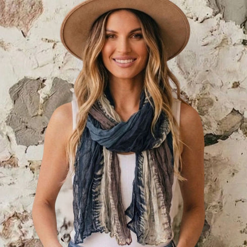 A woman wearing a wide-brimmed tan hat and a blue, gray, and beige patterned scarf, standing against a textured white and gray wall.