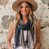 A woman wearing a wide-brimmed tan hat and a blue, gray, and beige patterned scarf, standing against a textured white and gray wall.