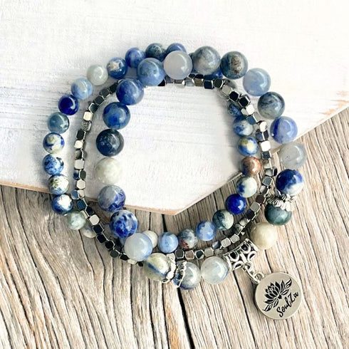 A stack of beaded stretch bracelets featuring blue, white, and gray gemstone beads, dark metallic spacer beads, and a silver-toned lotus flower charm engraved with the word 'Sukuna', displayed on a rustic wood surface next to a white hexagonal block.