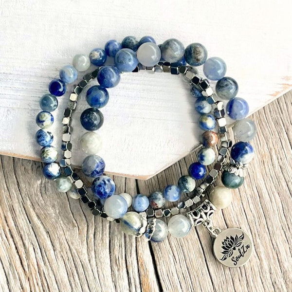 A stack of beaded stretch bracelets featuring blue, white, and gray gemstone beads, dark metallic spacer beads, and a silver-toned lotus flower charm engraved with the word 'Sukuna', displayed on a rustic wood surface next to a white hexagonal block.