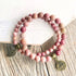 A beaded stretch bracelet made of round pink and mauve beads with two antique bronze charms, one leaf-shaped and one round lotus flower charm engraved with 'Love Me', displayed on a rustic wood surface next to a white wooden block.
