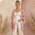 A woman stands against a textured beige wall with pampas grass props, wearing a lightweight open-front kimono cardigan with a pink and beige paisley floral print over a white tank top and white jeans.