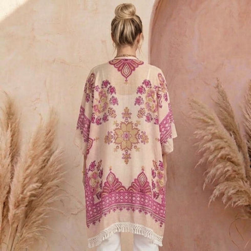 A woman viewed from behind wearing a loose pink kimono with pink and gold floral embroidery, standing in front of a textured pink wall with pampas grass decor.