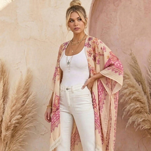 A woman models a lightweight beige kimono-style cardigan with pink and orange floral patterns, worn open over a white tank top and white jeans, set against a warm textured beige wall with pampas grass.