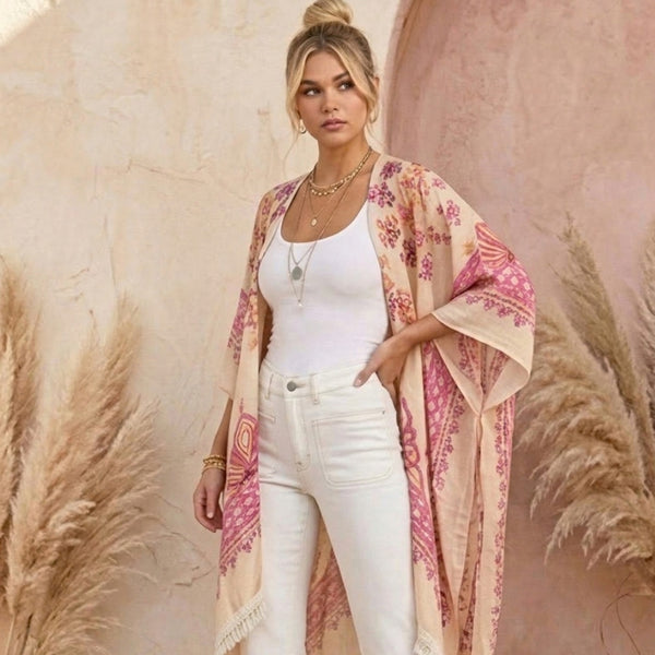 A woman models a lightweight beige kimono-style cardigan with pink and orange floral patterns, worn open over a white tank top and white jeans, set against a warm textured beige wall with pampas grass.