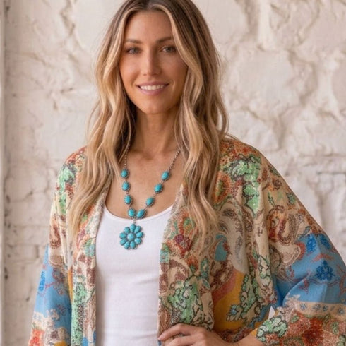 A woman with long blonde hair wearing a white tank top, a turquoise flower necklace, and a lightweight multicolor paisley cardigan, standing against a textured white wall.