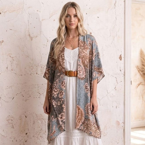 A woman stands indoors wearing a loose, patterned open-front Soulza Boho Kimono Cardigan over a white boho dress, with a brown belt and tan sandals. The room has a textured cream wall and a doorway with dried grass decor.