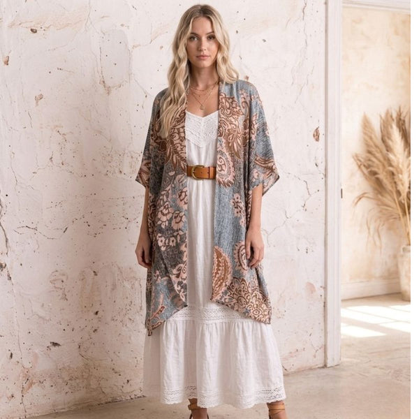 A woman stands indoors wearing a loose, patterned open-front Soulza Boho Kimono Cardigan over a white boho dress, with a brown belt and tan sandals. The room has a textured cream wall and a doorway with dried grass decor.